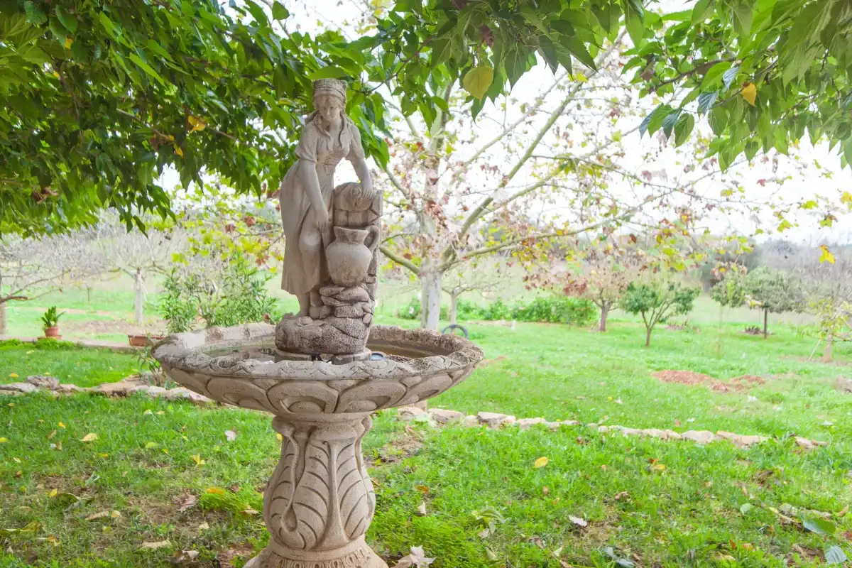 A garden fountain stands beneath leafy trees in a grassy orchard.