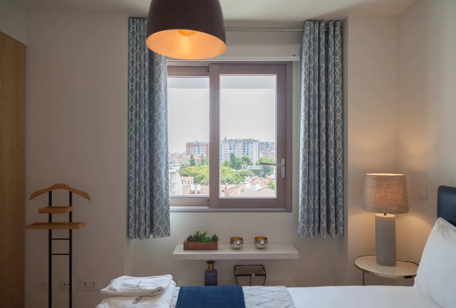 A bedroom with a window overlooking nearby buildings and trees.