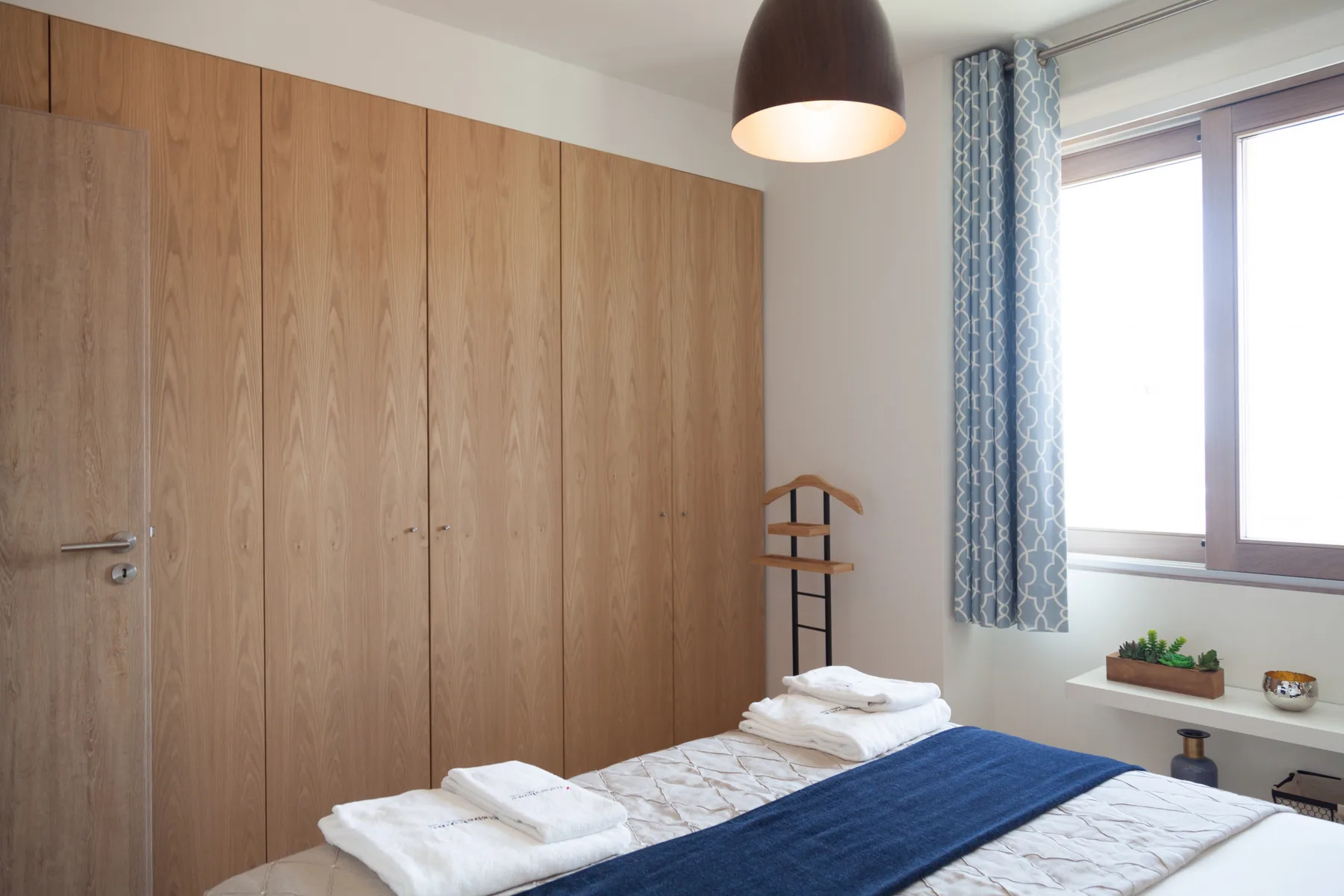 A bedroom with a bed, folded towels, and a window with blue curtains.
