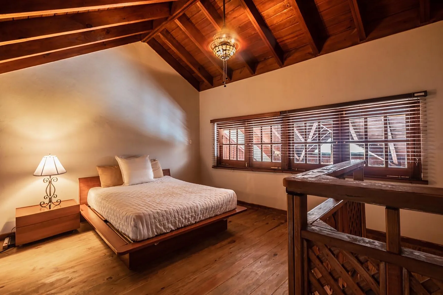 A bedroom with a bed beneath a vaulted wooden ceiling and windows with blinds.