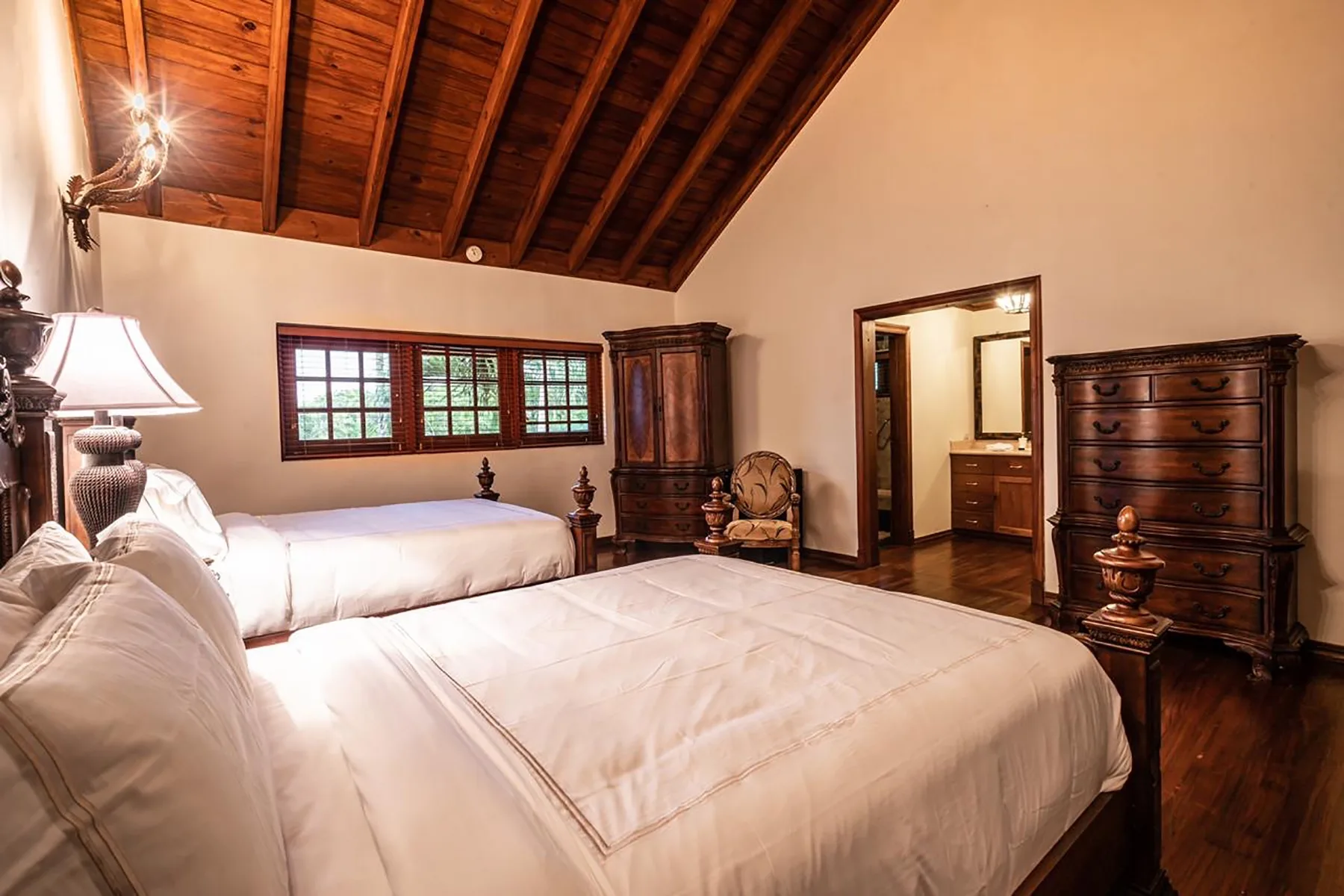 A bedroom with white bedding, dark wooden furnishings, and a sloped timber ceiling.