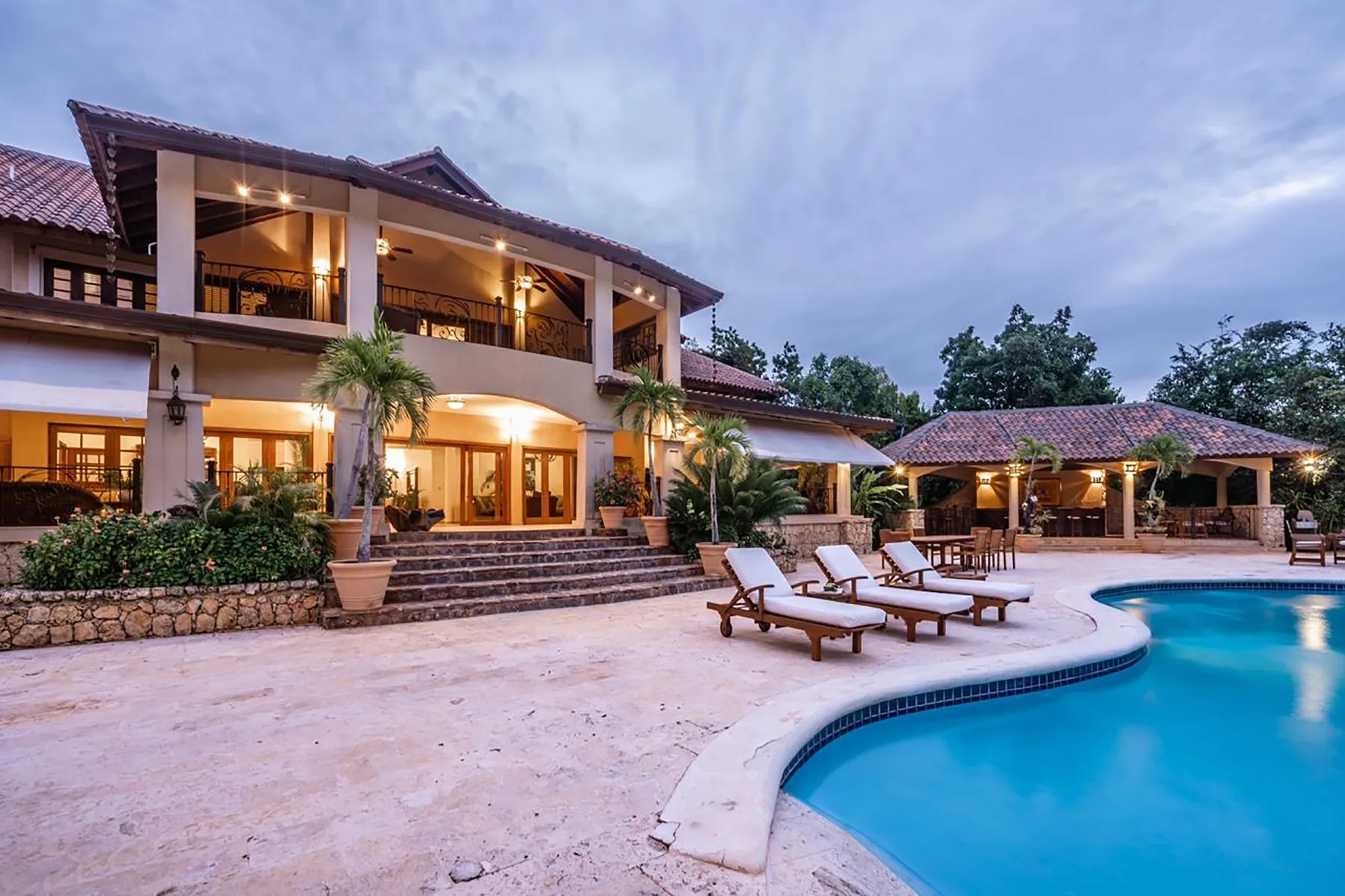 A large house opens onto a curved swimming pool and a covered outdoor seating area.