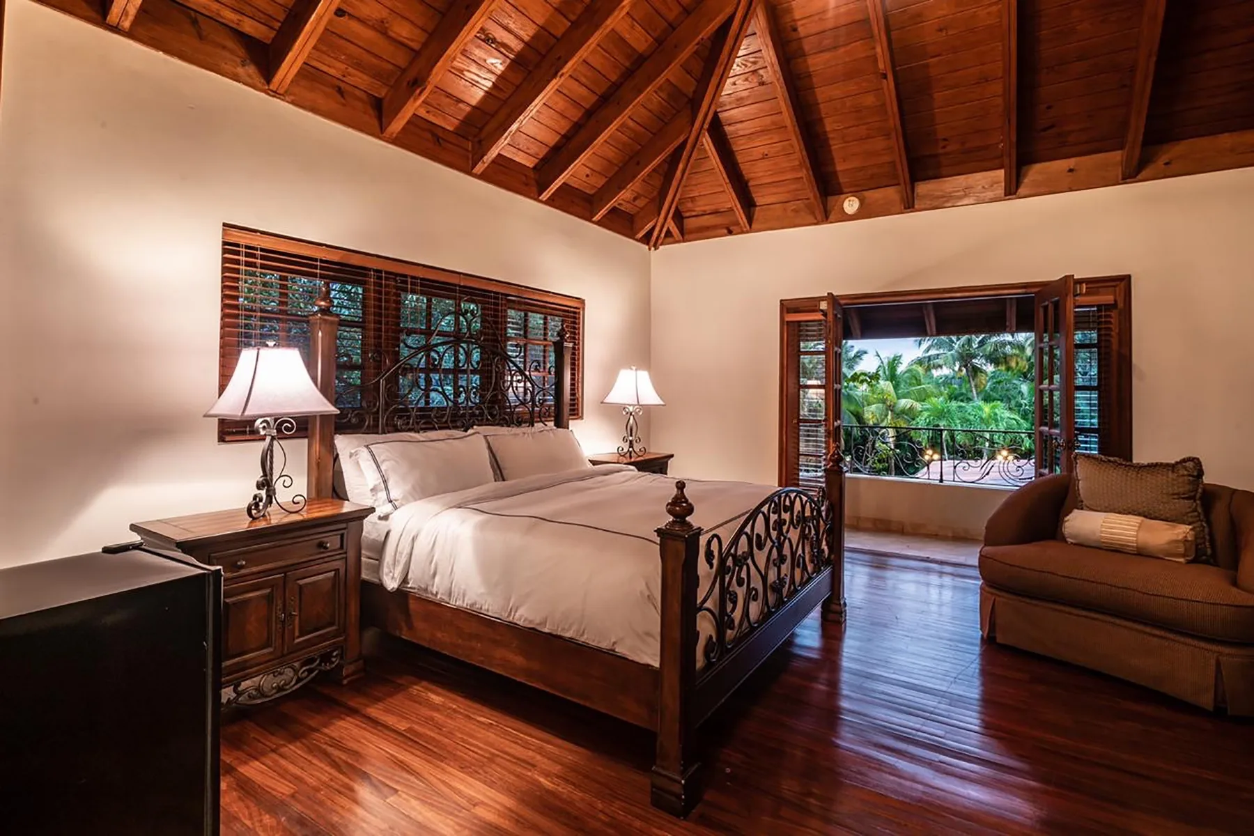 A bedroom with a large bed, wooden floors, and windows overlooking greenery.