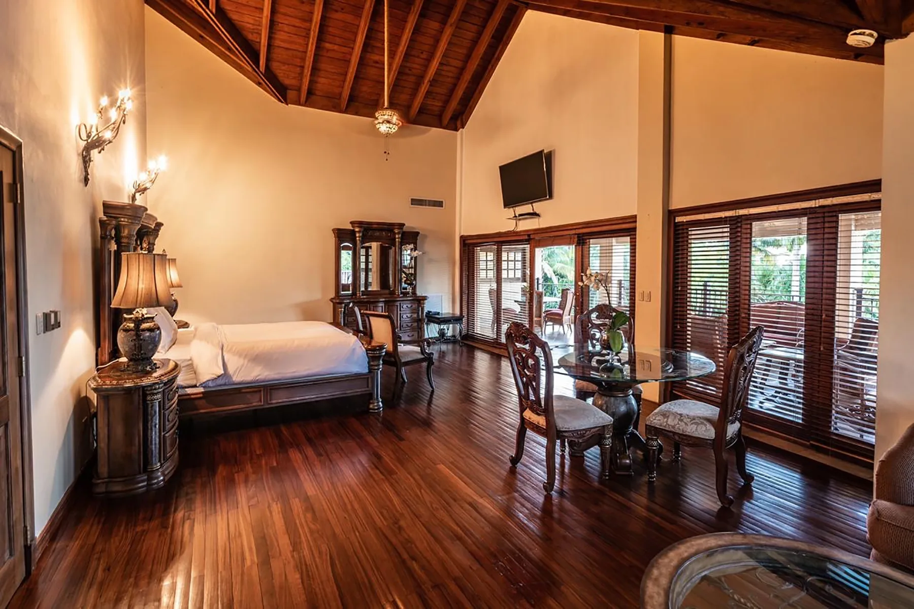 A bedroom with a bed, dining table, and large windows beneath a high wooden ceiling.