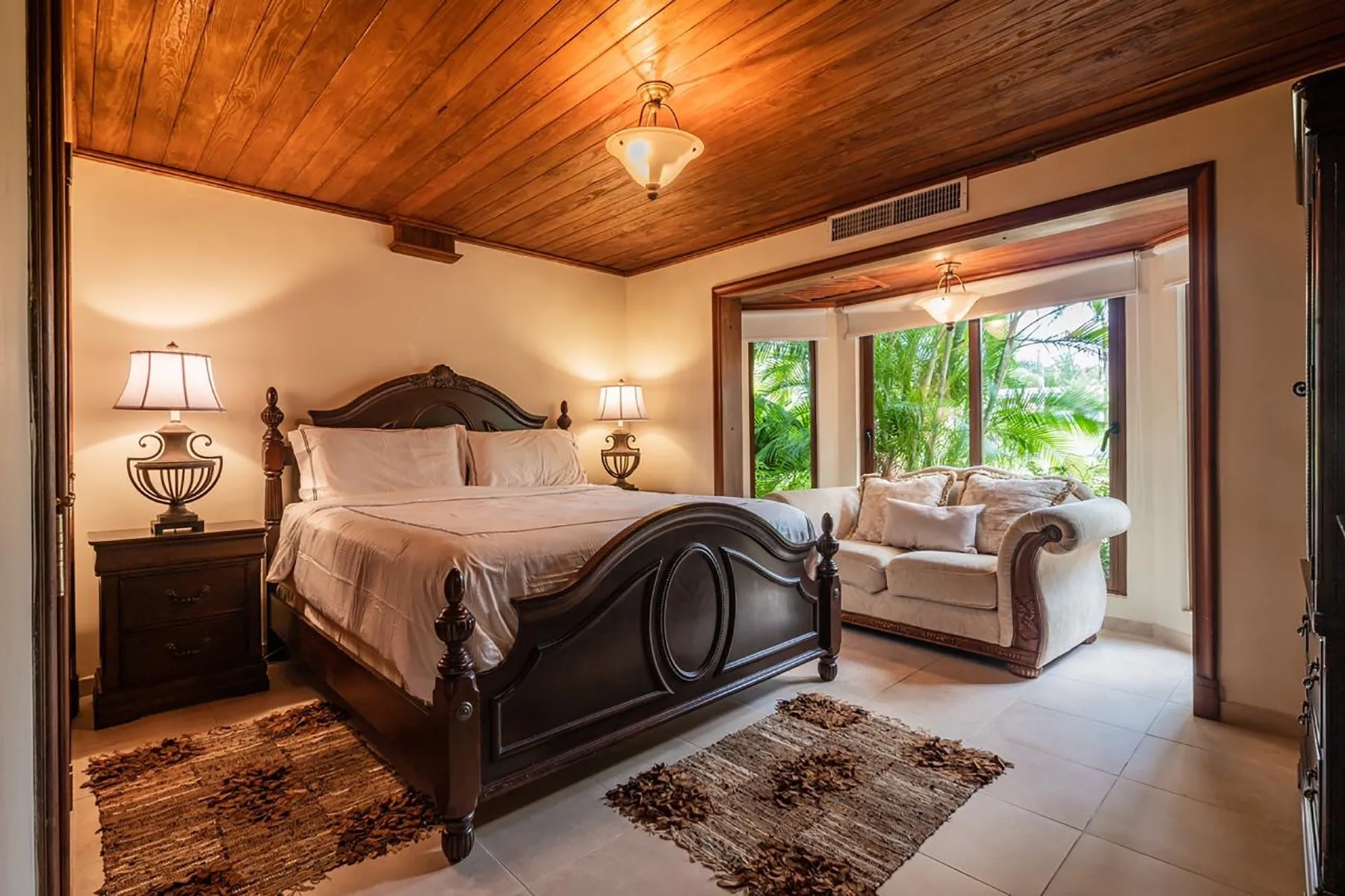 A bedroom with a large bed and a seating area beside bright windows.
