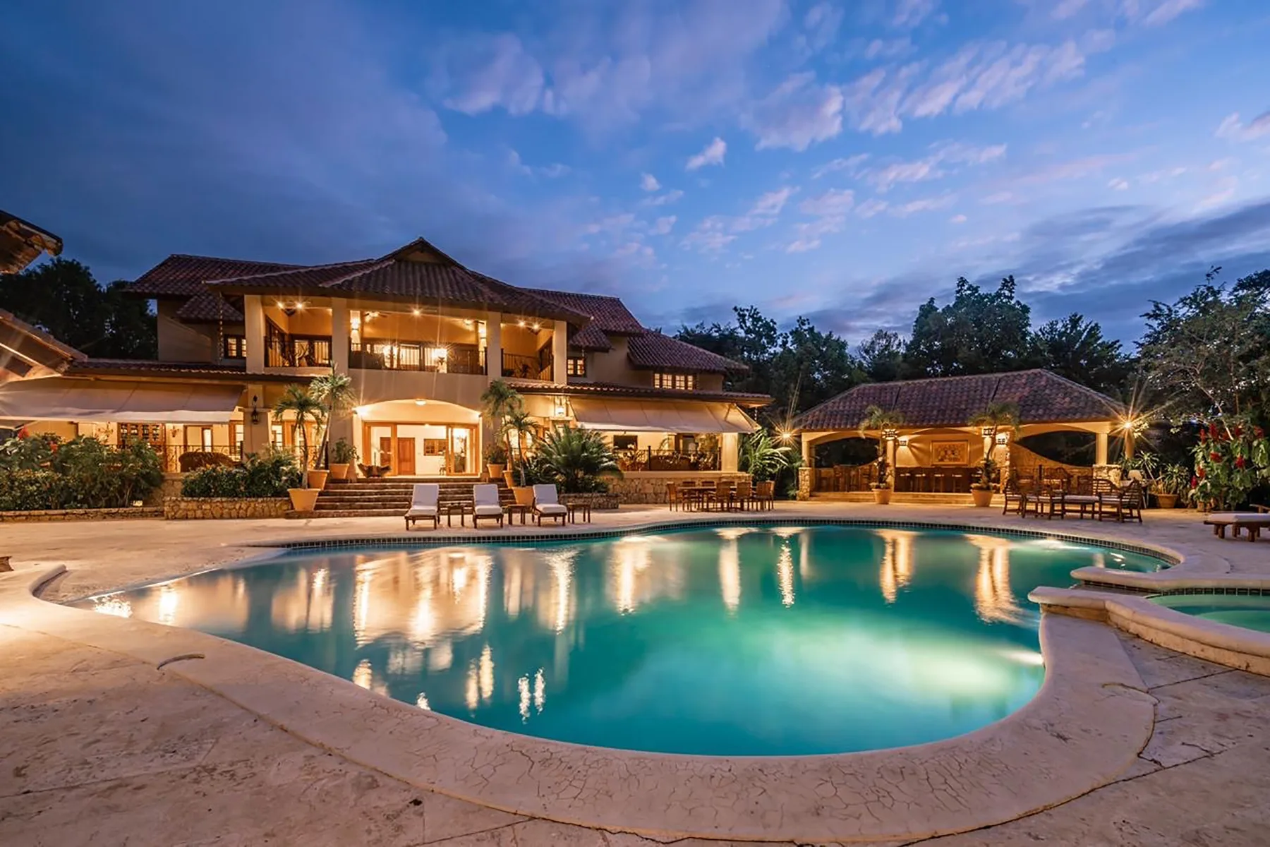 An outdoor swimming pool glows beside a warmly lit building at dusk.