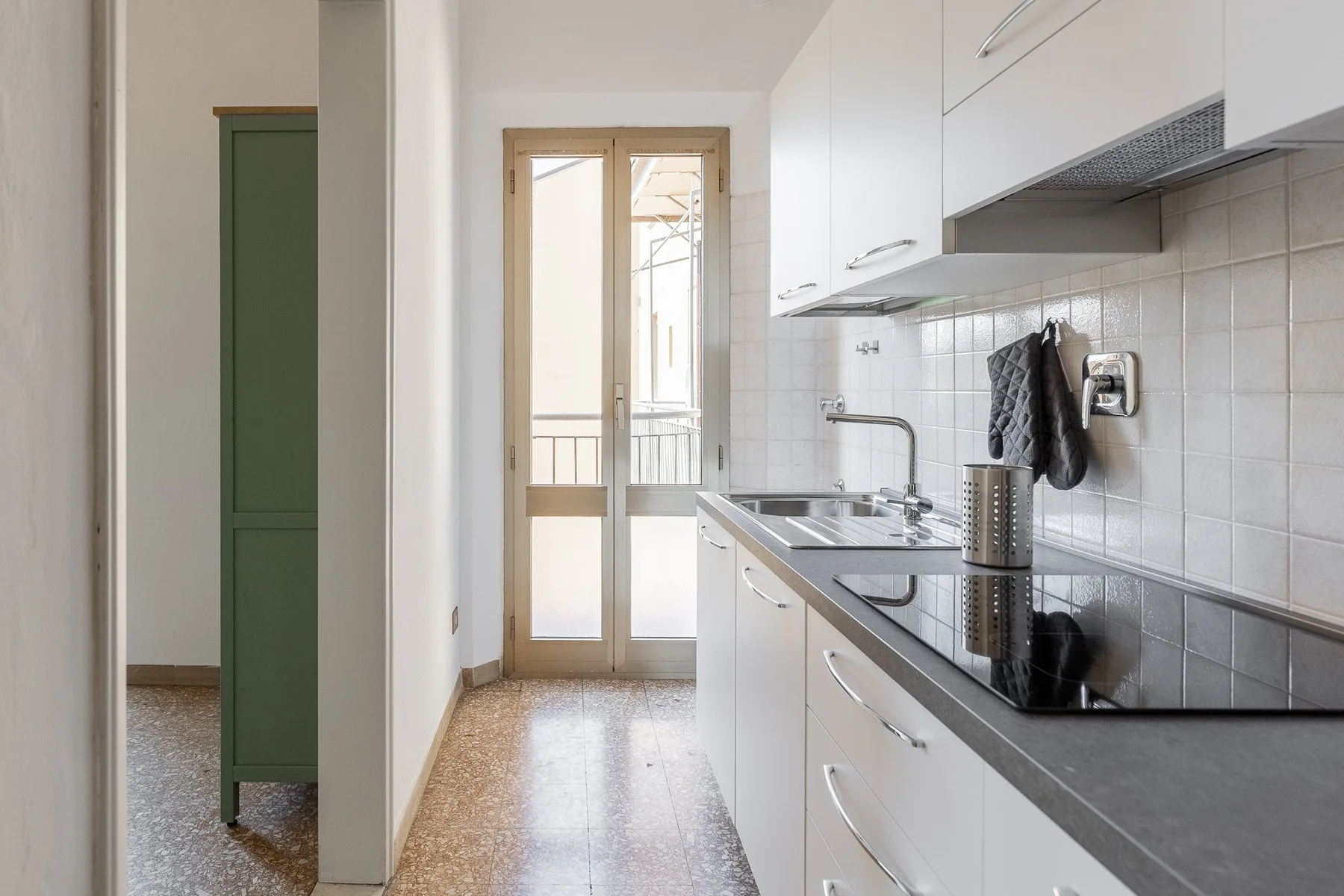 A narrow kitchen with white cabinets, a dark countertop, and a glass door at the far end.