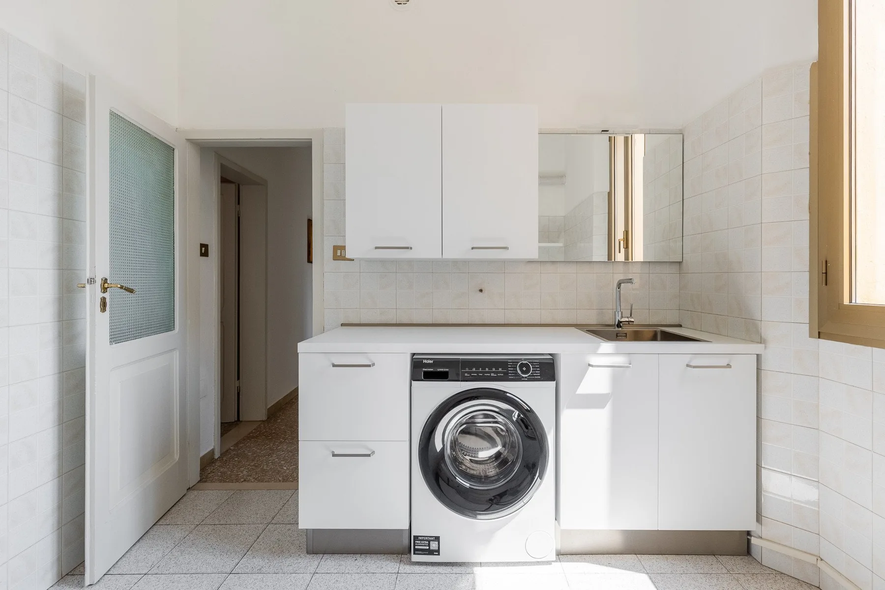 A bright tiled kitchen with white cabinets, a washing machine, and a sink.