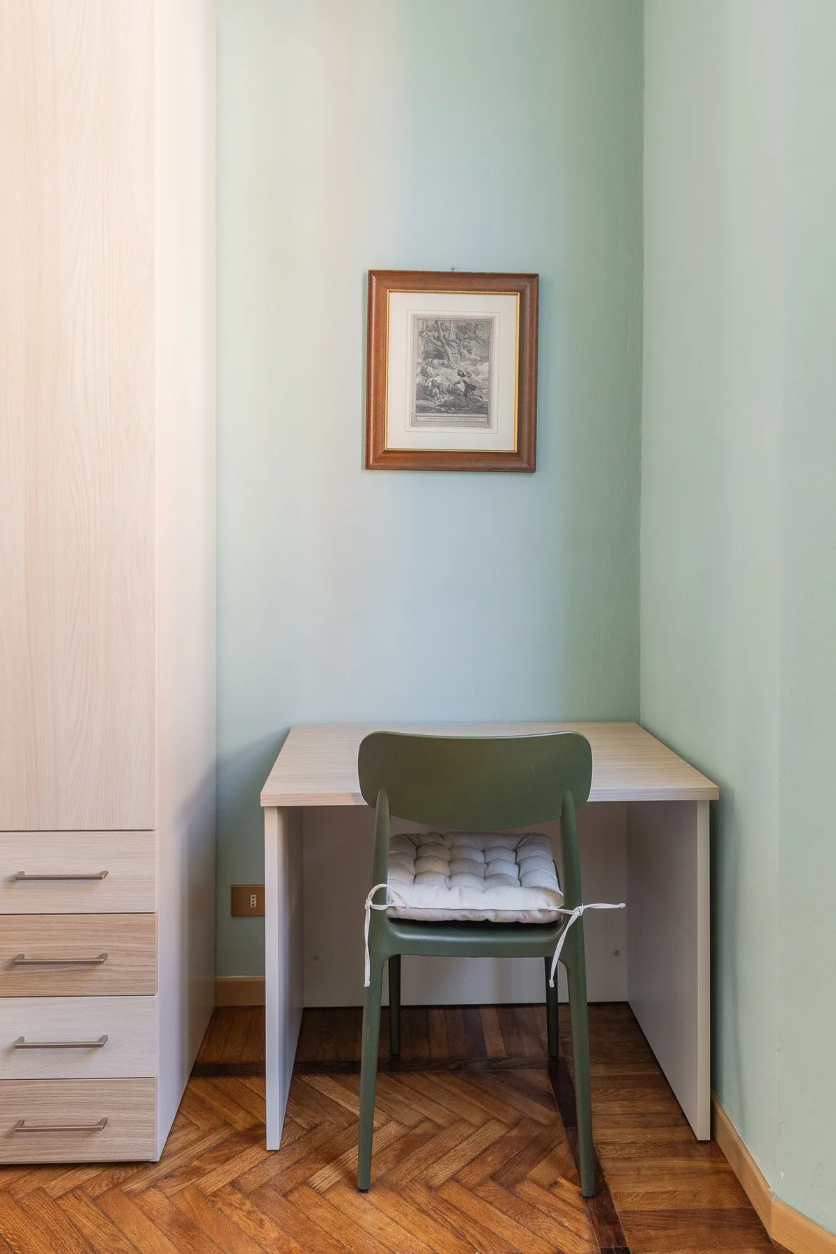 A compact desk and chair sit against a pale green wall beneath framed artwork.