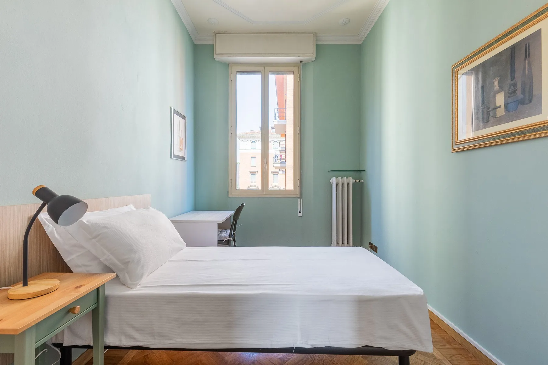 A bedroom with a neatly made bed, a desk, and a tall window.