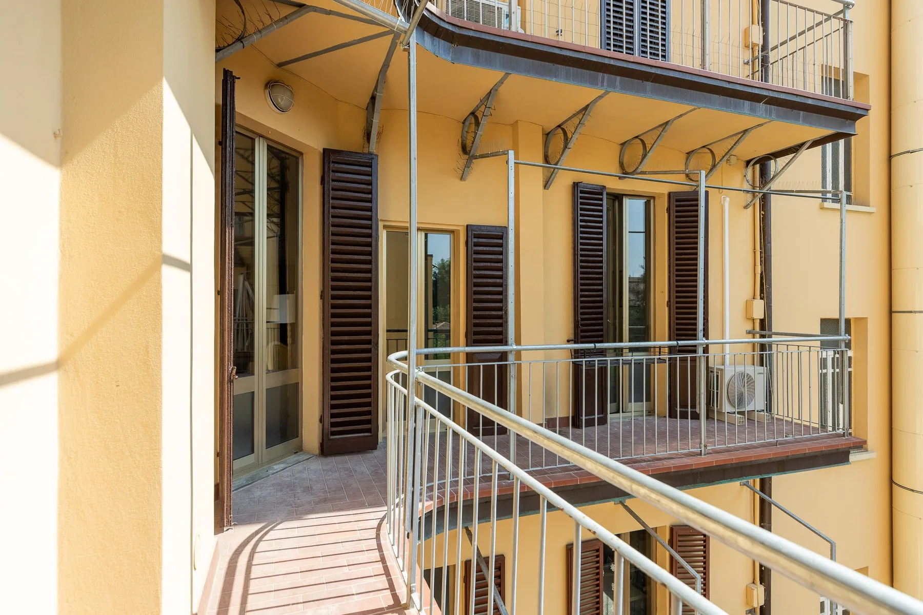 A sunlit exterior balcony with metal railings and shuttered windows.