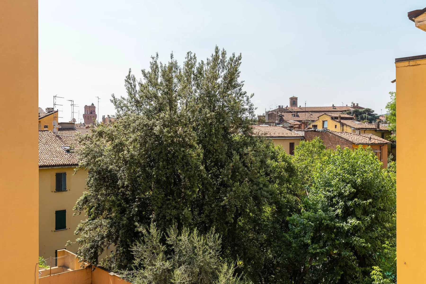 A leafy outdoor view overlooks trees and tiled rooftops beneath a clear sky.