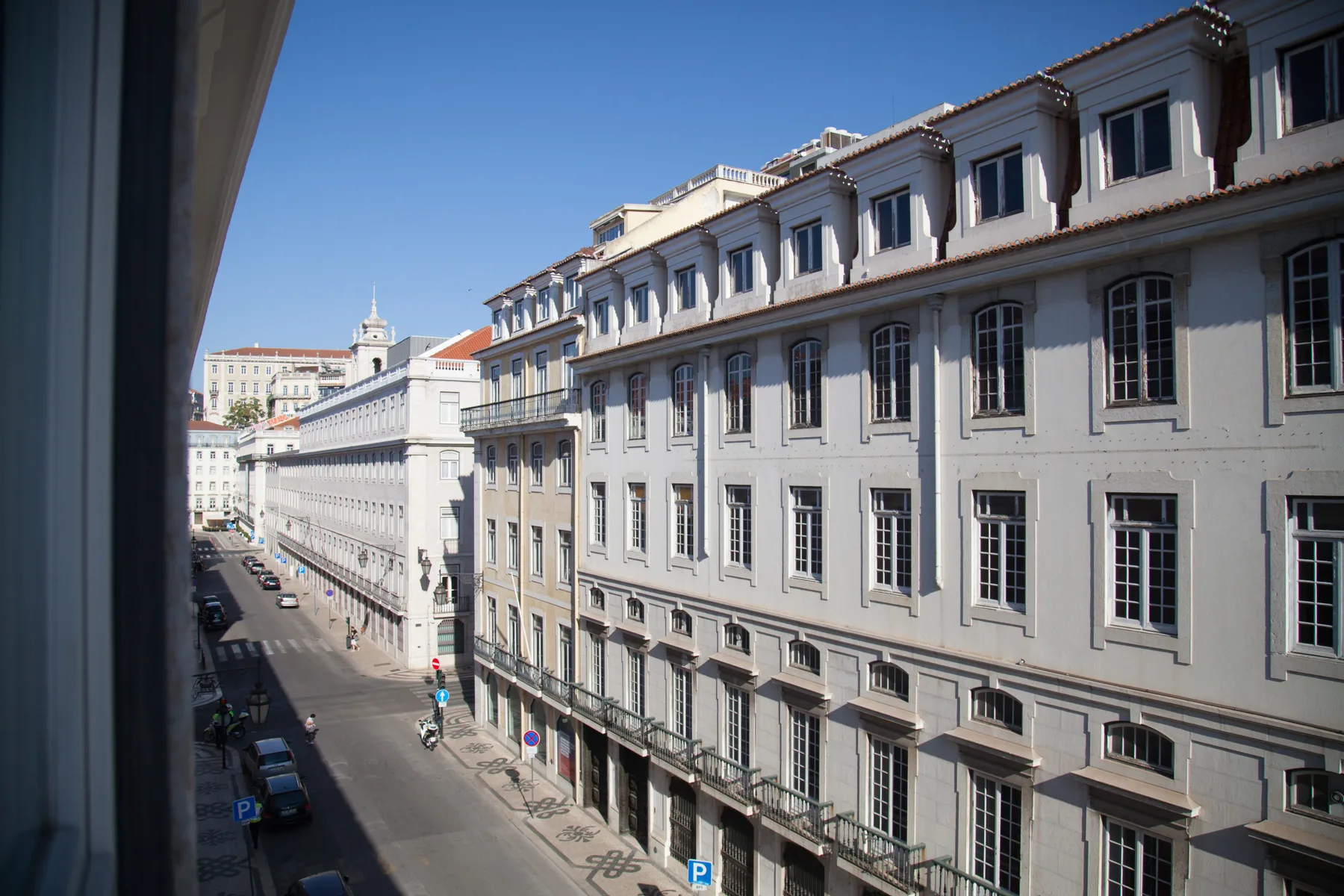 Una calle urbana bordea una hilera de edificios claros bajo un cielo despejado.