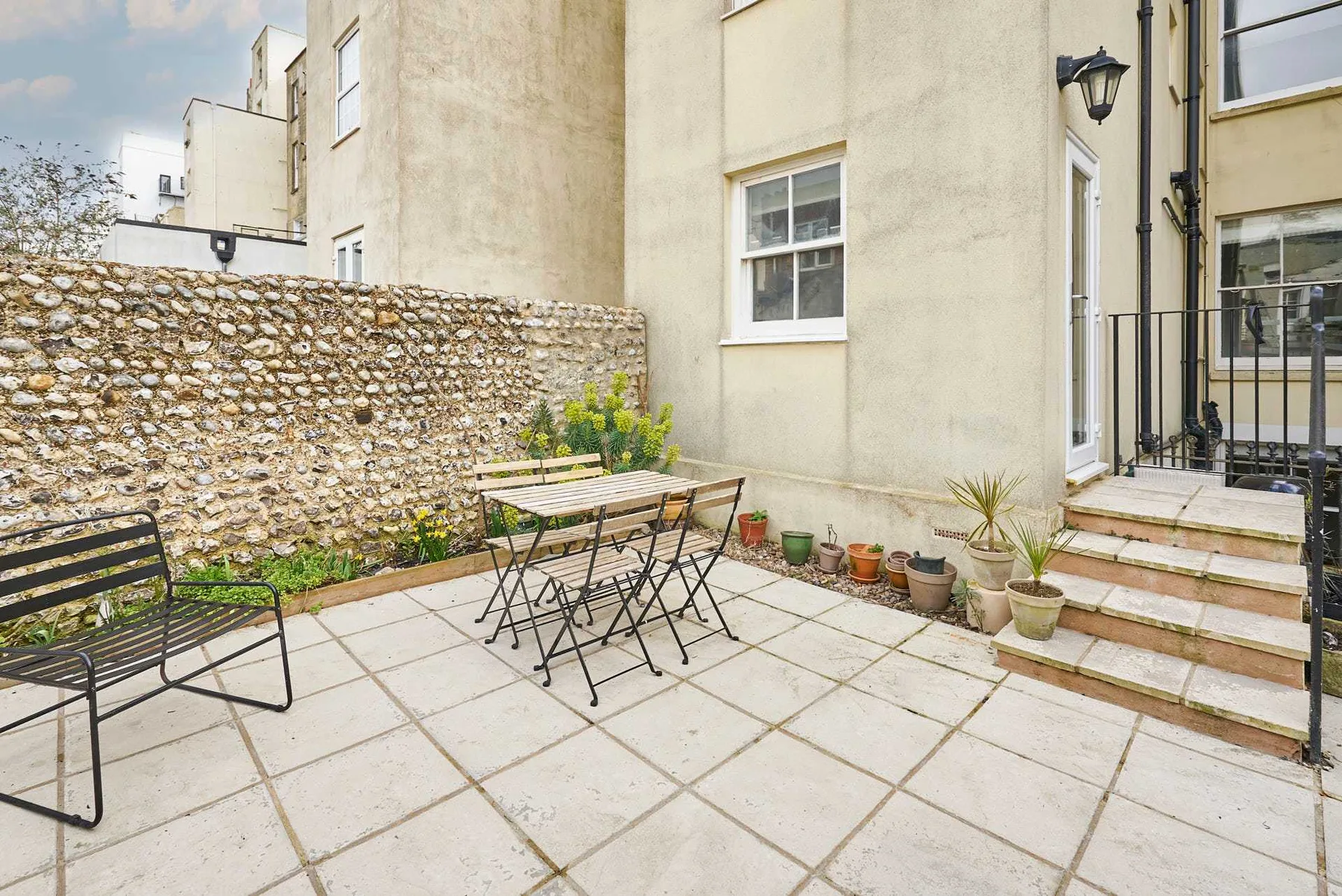 A paved outdoor patio with a table, seating, potted plants, and a stone wall.