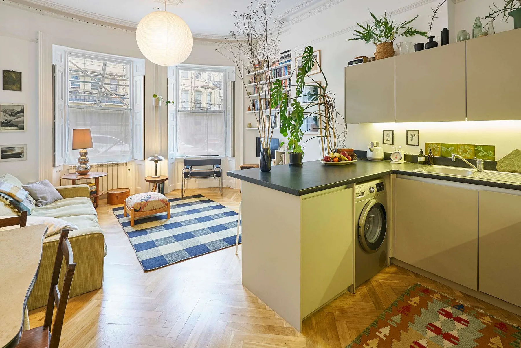 A bright open-plan living room and kitchen with large windows and a blue checked rug.