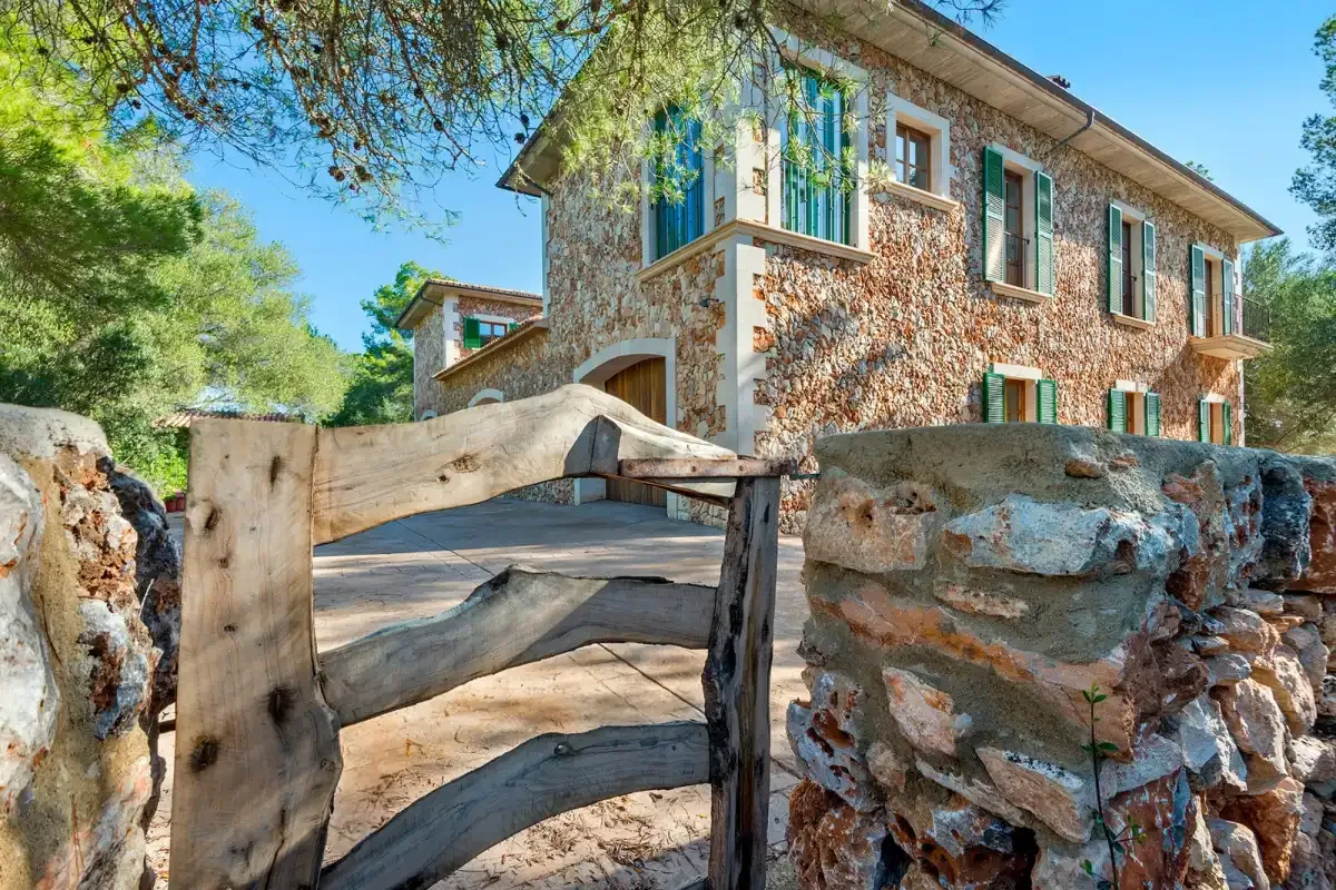 A stone house stands behind a rustic wooden gate and low stone wall.