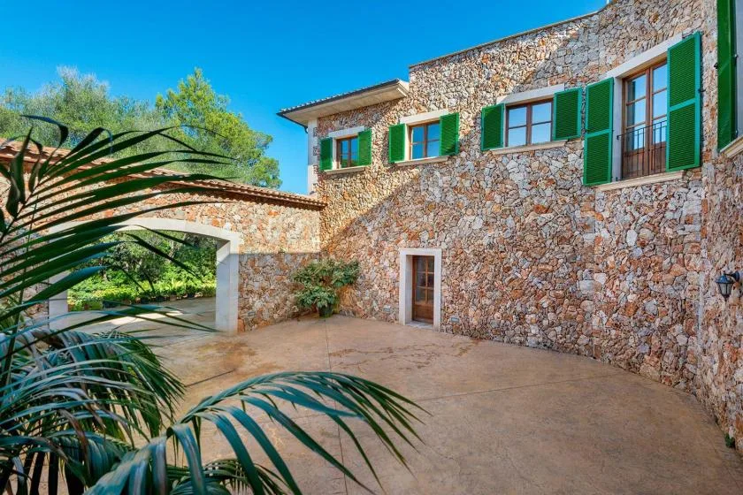 A stone house opens onto a paved courtyard, with green shutters along the upper windows.