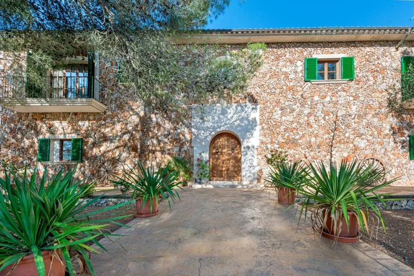 A stone house entrance opens onto a paved courtyard with potted plants.