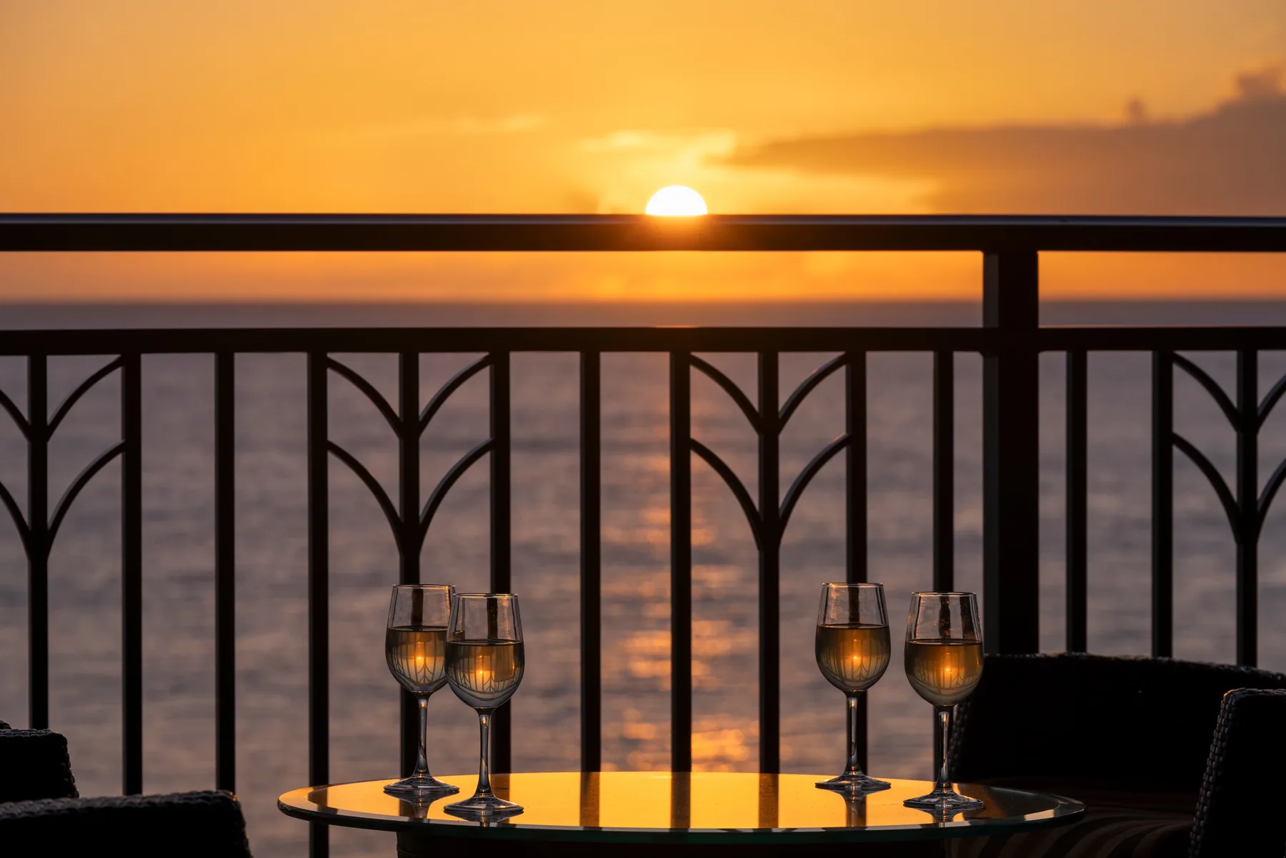 Una mesa de balcón con copas da al agua al atardecer.