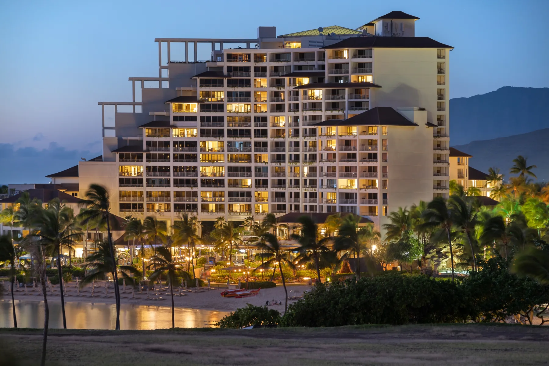 Un gran edificio turístico se ilumina al atardecer detrás de palmeras y jardines.