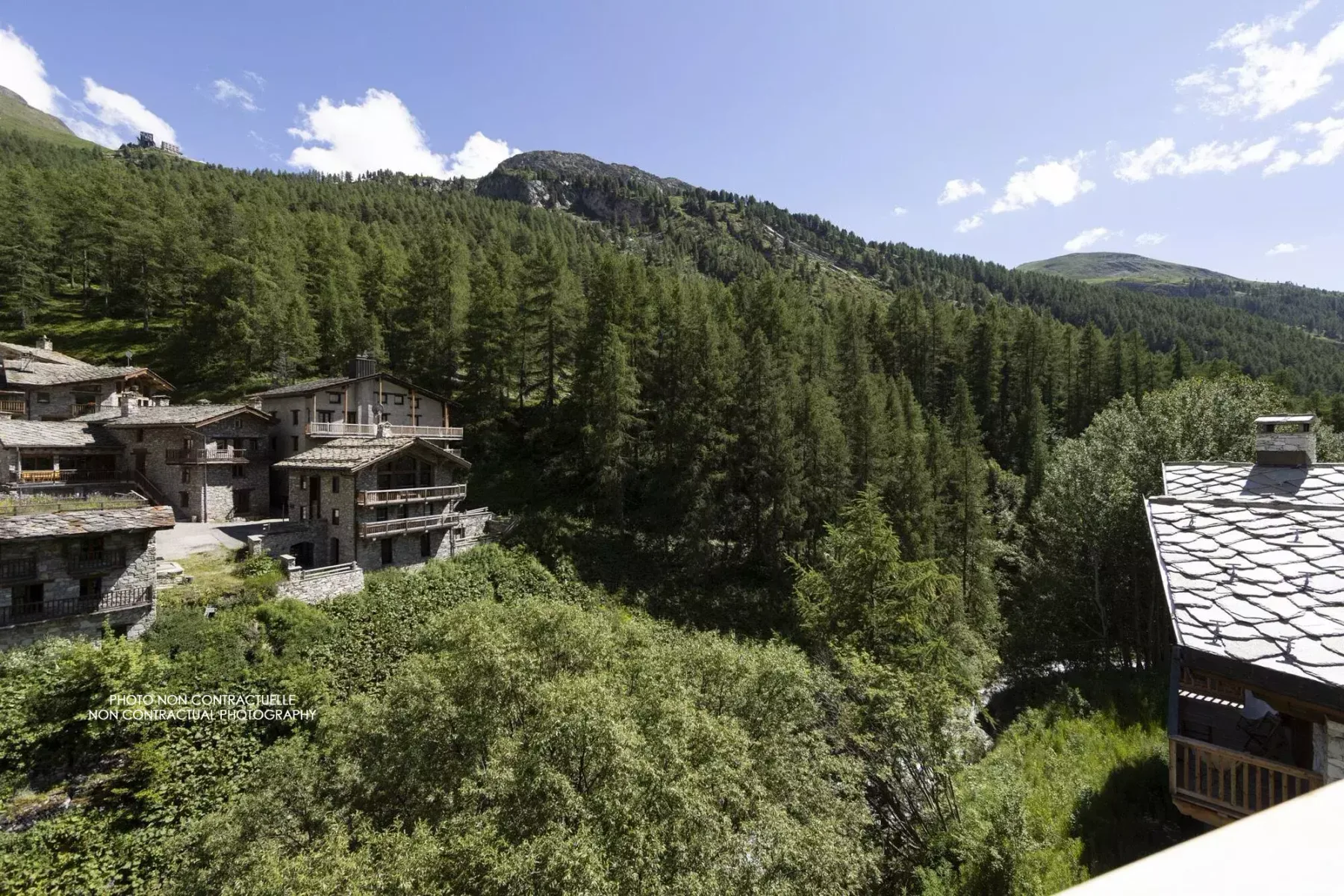 Mountain village buildings sit among dense forest beneath a clear, cloud-dotted sky.