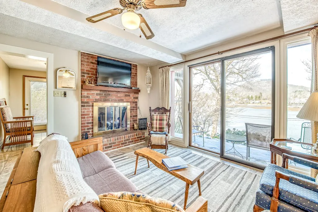 A living room with a brick fireplace, seating, and large windows overlooking the water.