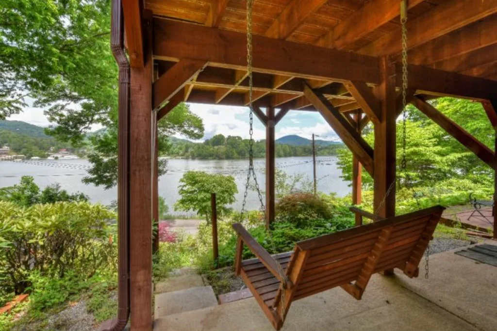 A covered porch overlooks a lake, with a wooden swing hanging beneath the roof.