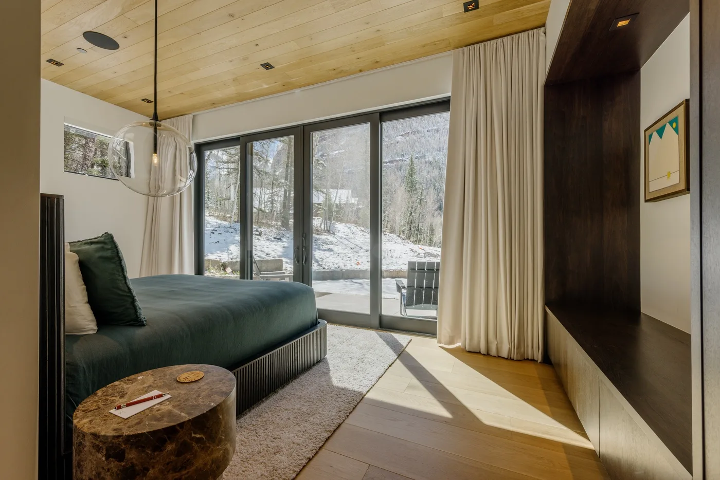 Dormitorio con una cama junto a grandes ventanas con vistas a un paisaje boscoso.