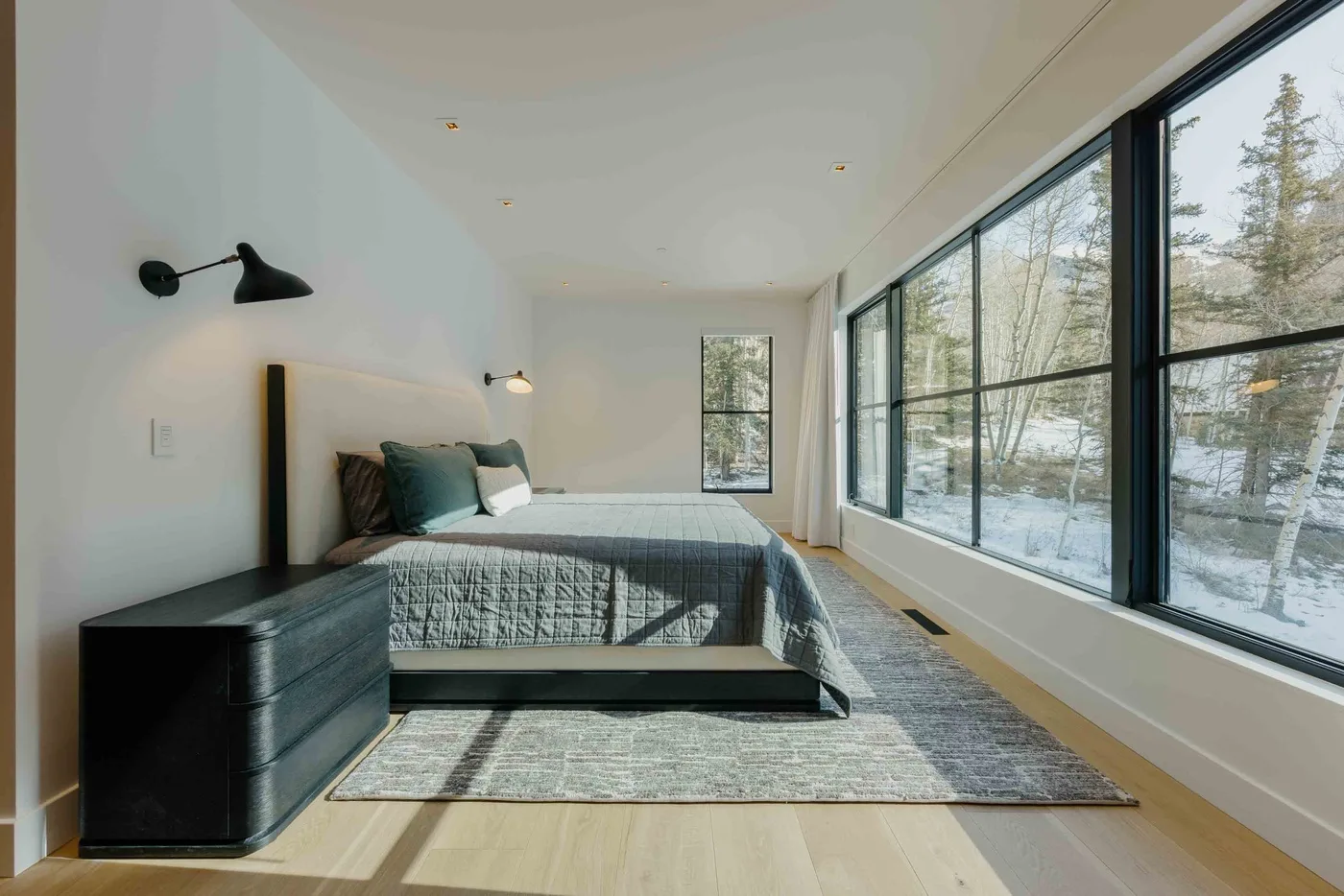 Un dormitorio luminoso con una cama junto a grandes ventanas con vistas a los árboles.