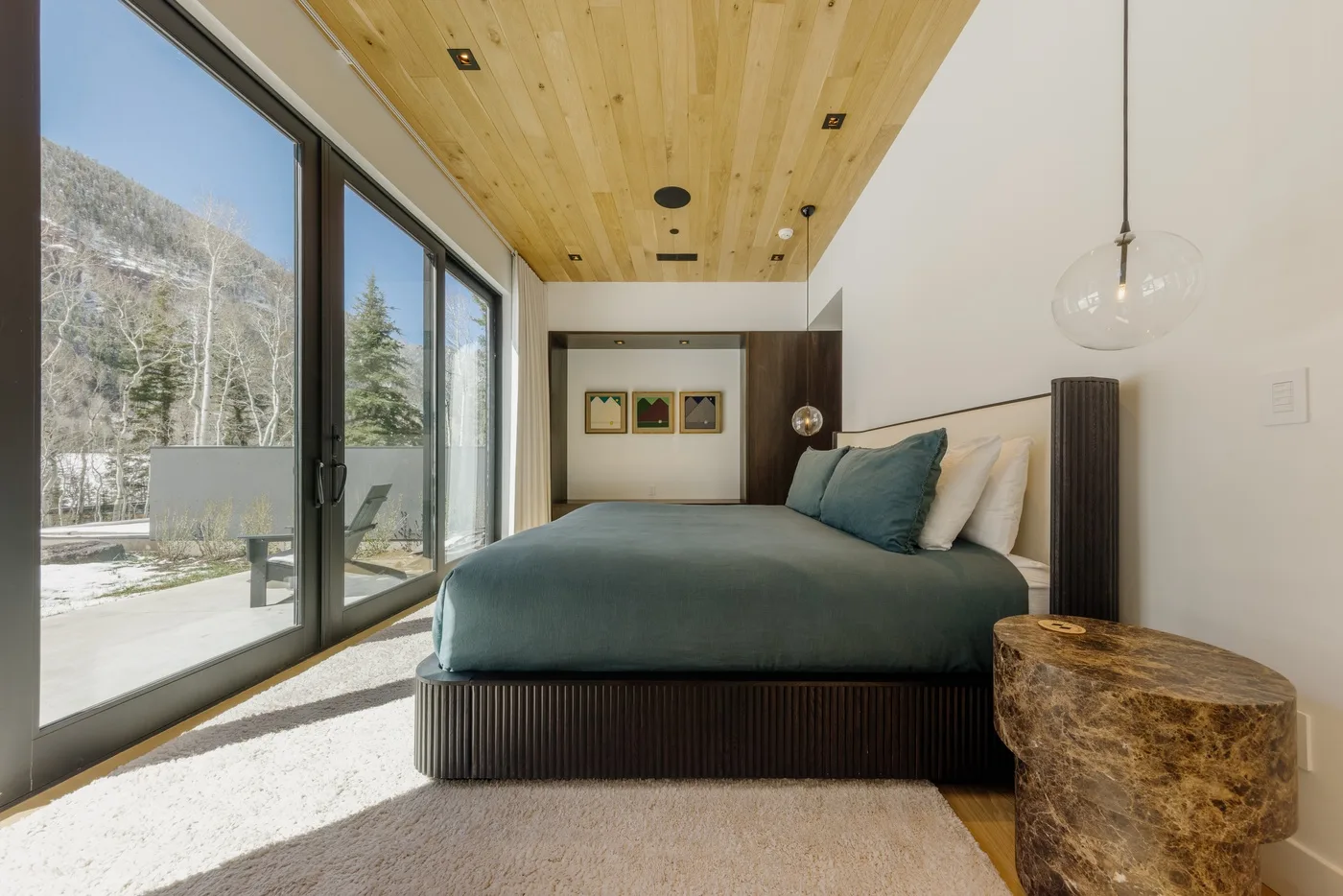 Un dormitorio con una cama, ropa de cama verde azulado y grandes ventanas con vistas a los árboles.