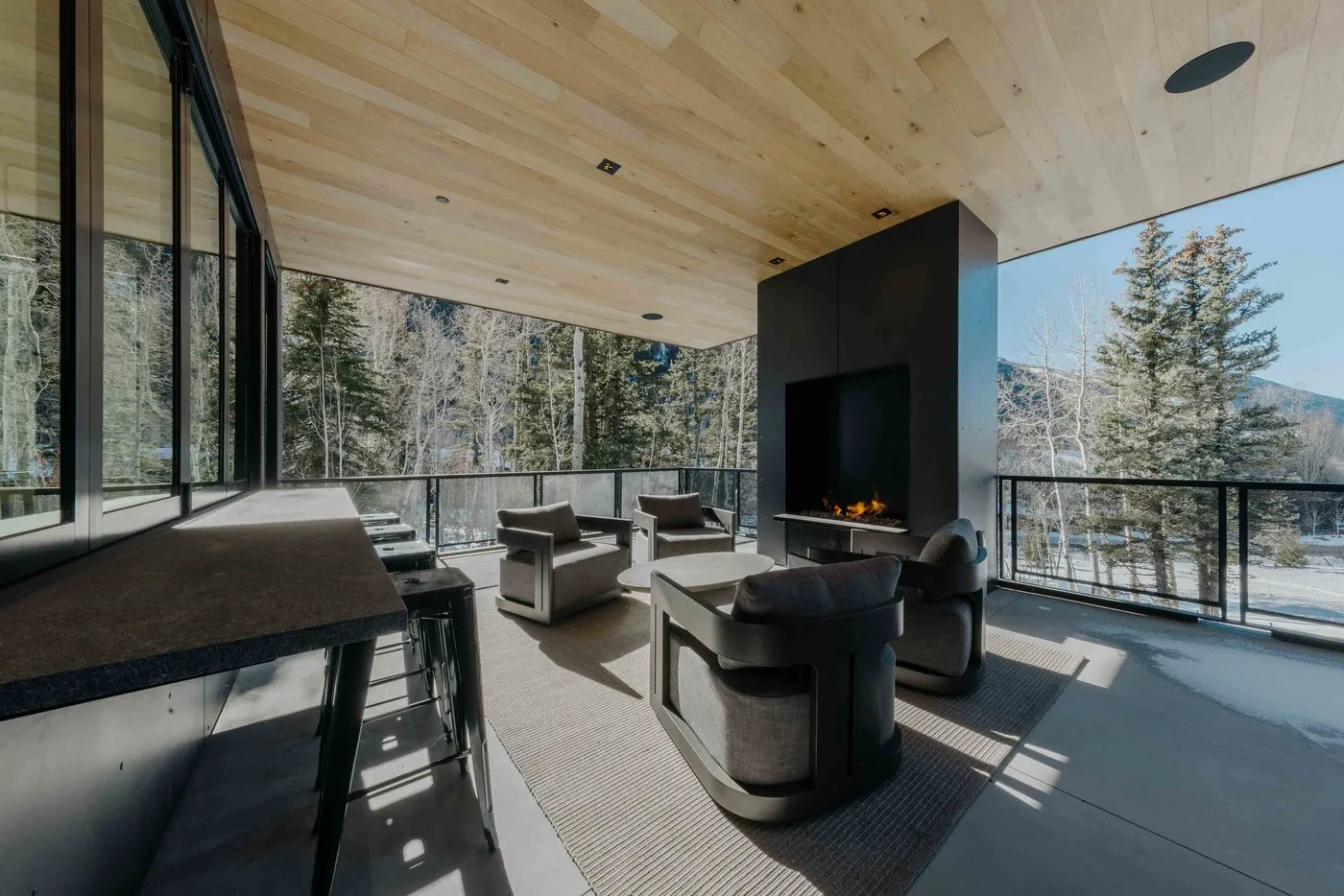 Una sala moderna con asientos alrededor de una chimenea y grandes ventanales con vistas a los árboles.