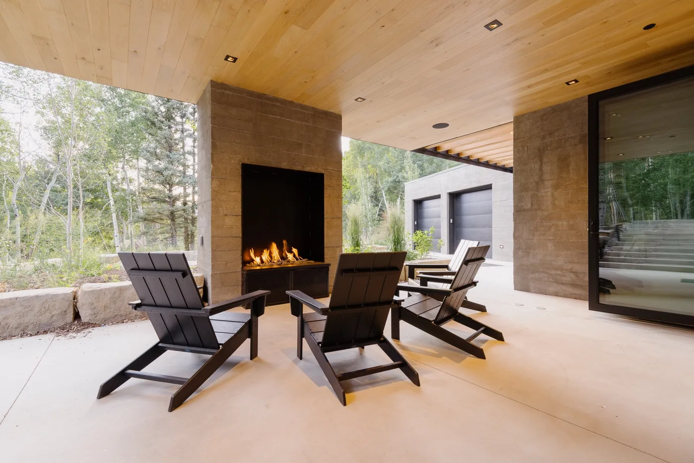 Una zona de estar exterior cubierta da a una chimenea encendida y vistas arboladas.