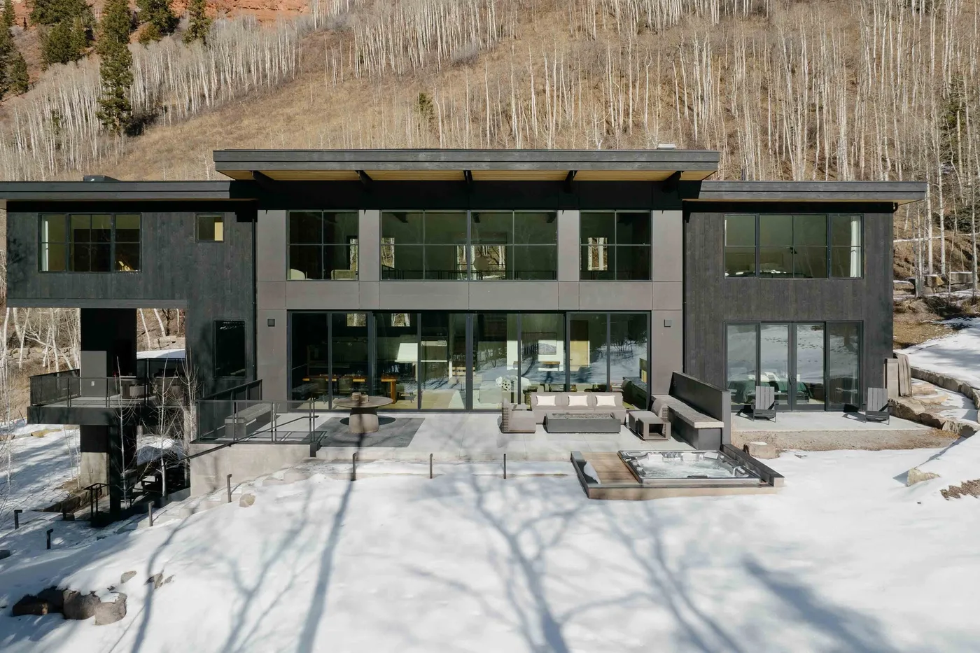 Una casa moderna con amplios ventanales se alza frente a una ladera boscosa y nevada.