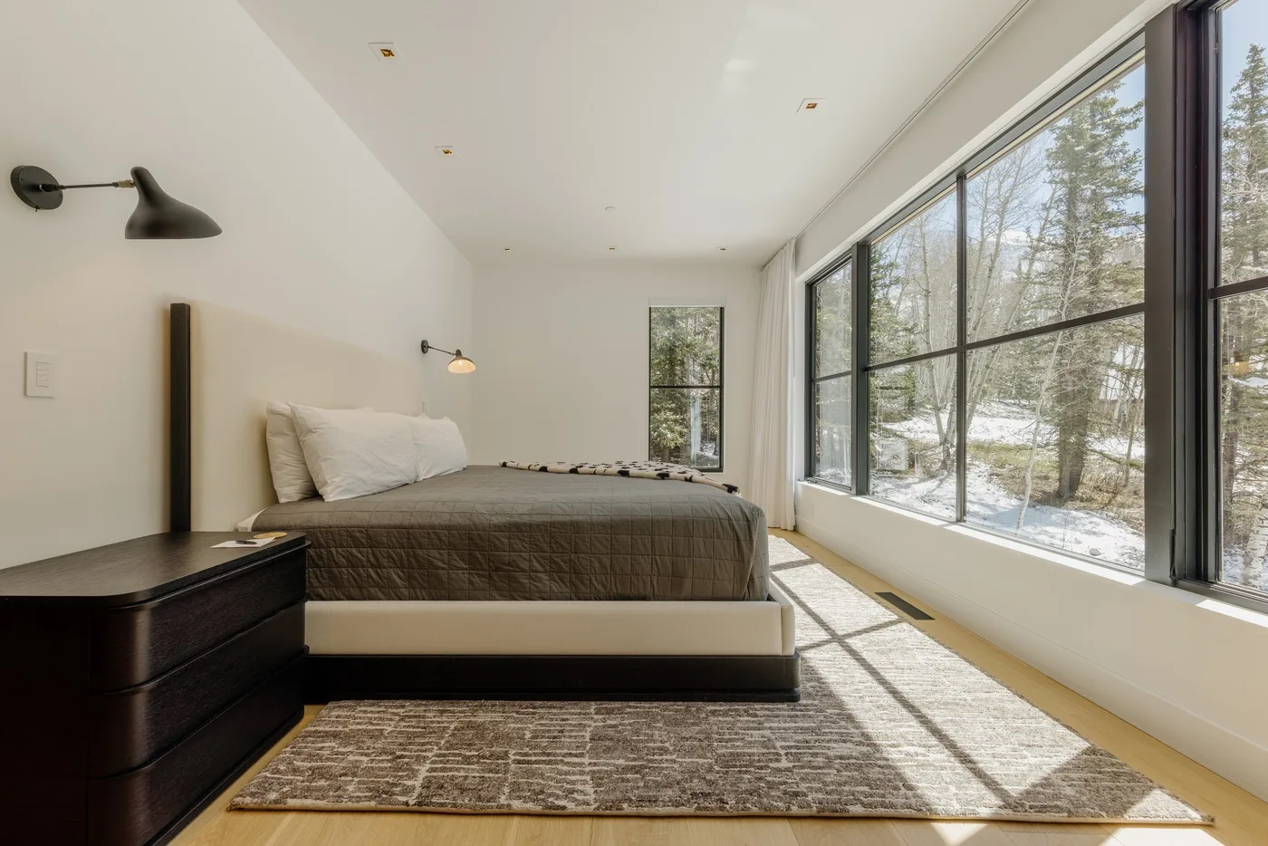 Un dormitorio luminoso con una cama junto a grandes ventanas que dan a los árboles.