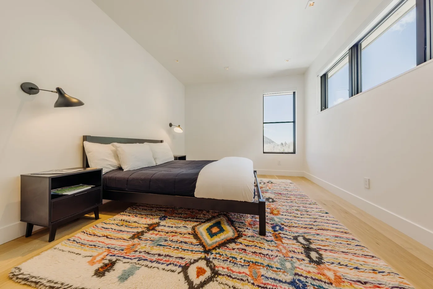 Un dormitorio con una cama baja, ropa de cama oscura y una alfombra colorida estampada.
