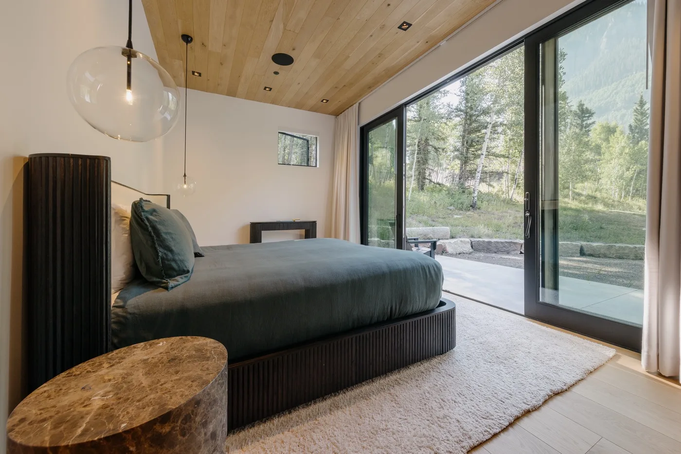 Un dormitorio con una cama junto a grandes puertas de vidrio con vista a los árboles.