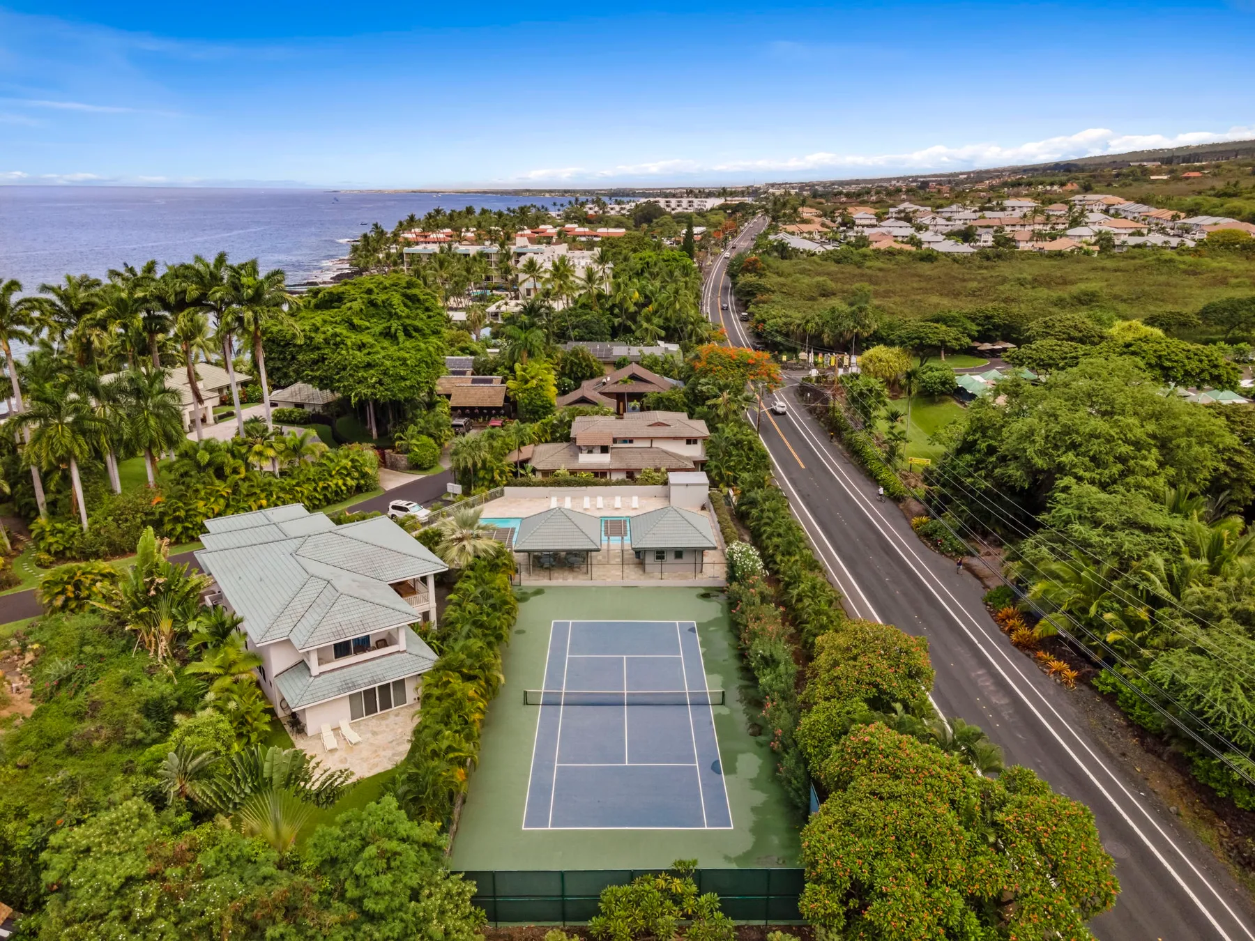 Vista aérea de un barrio costero con canchas de tenis, vegetación y una carretera curva.