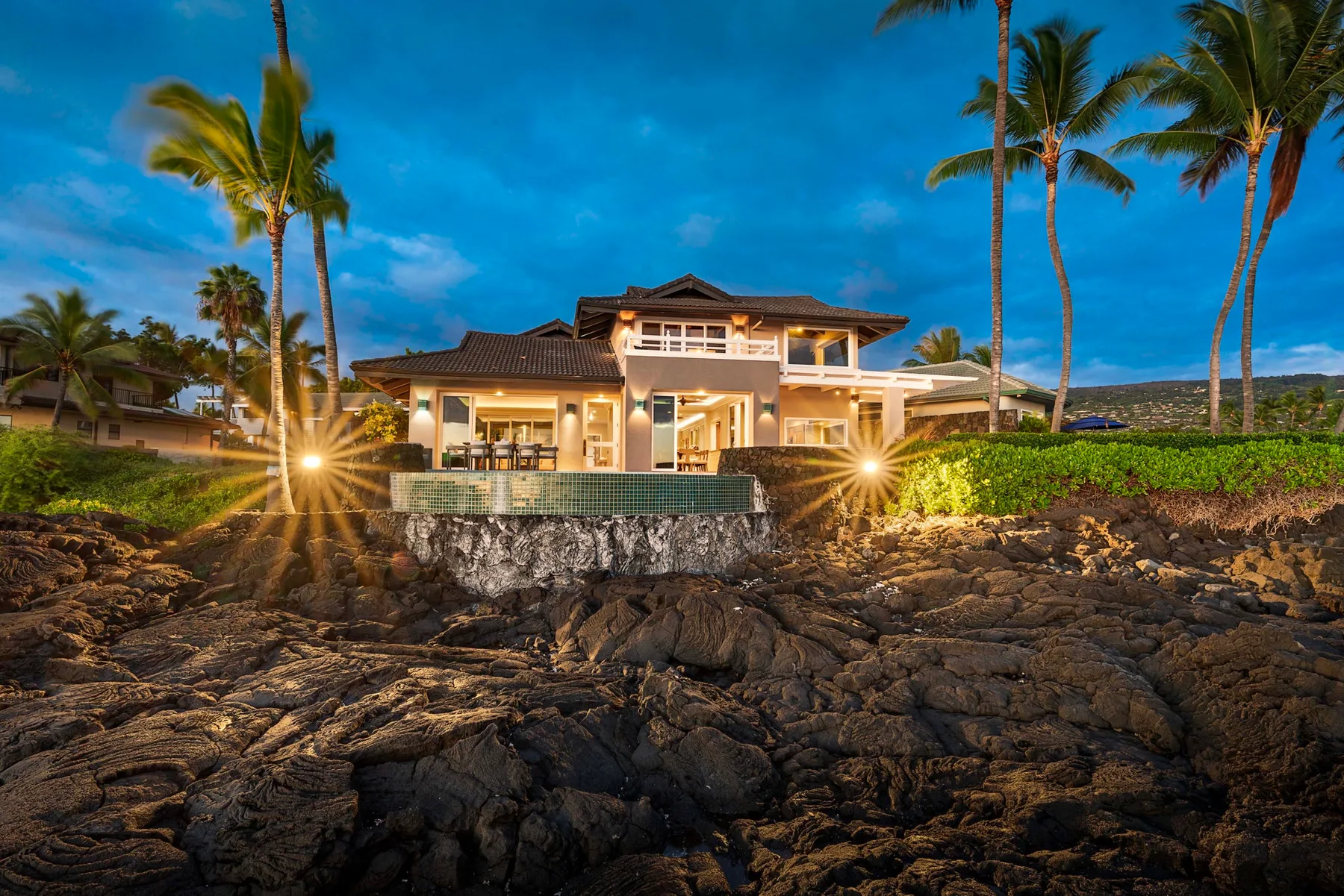 Una casa iluminada junto a la costa se alza detrás de una orilla rocosa bajo palmeras.