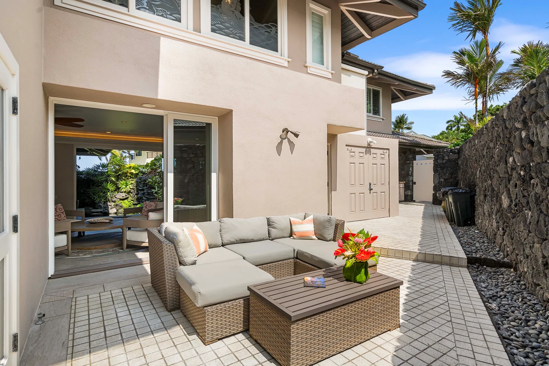 Un patio exterior tiene asientos acolchados, una mesa baja y plantas con flores.