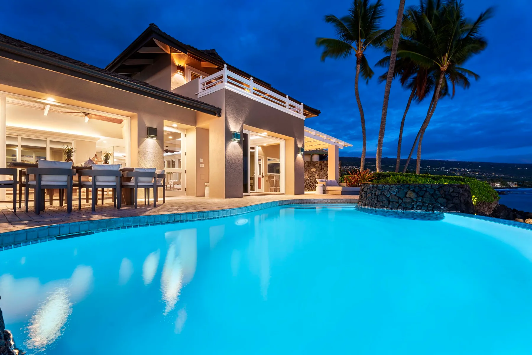 Una casa y una piscina exterior iluminadas junto a palmeras al anochecer.
