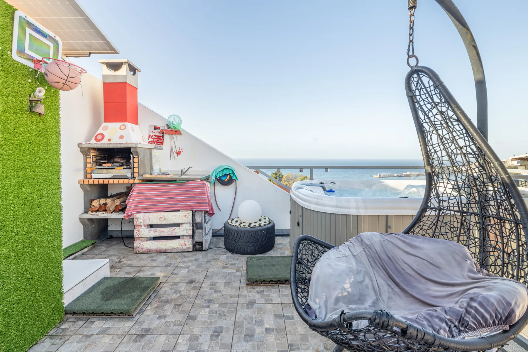 A tiled outdoor terrace has a barbecue, hot tub, and hanging seat overlooking the sea.