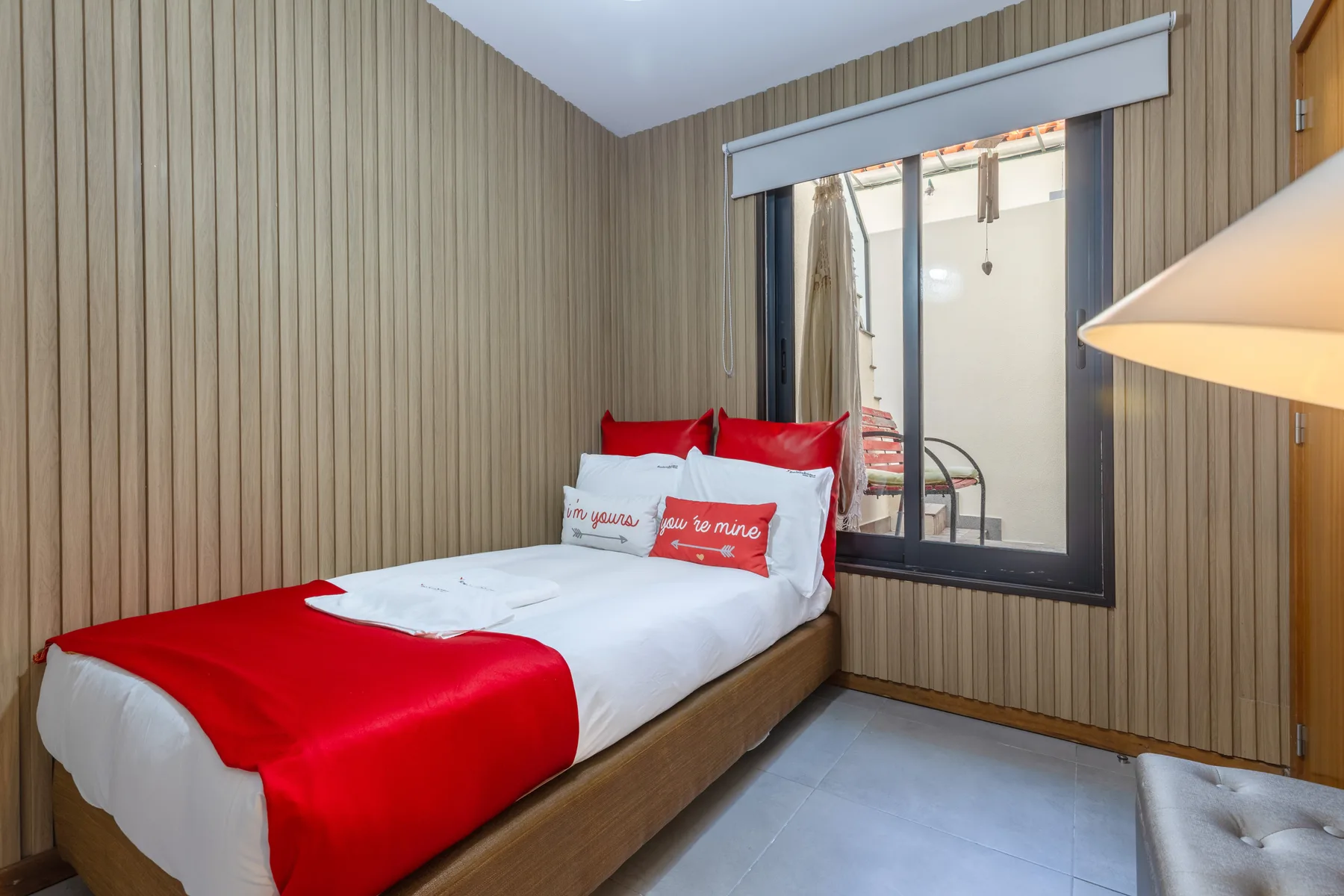 A bedroom with a bed dressed in white and red beside a large window.