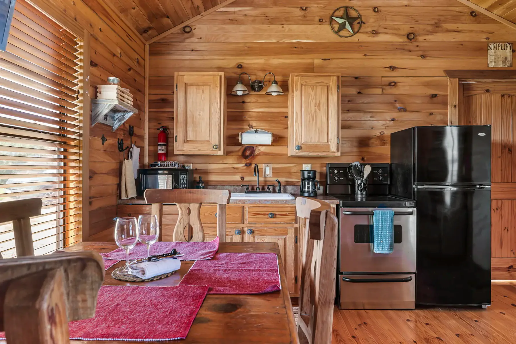 Acogedora cocina de cabaña de madera con comedor, refrigerador negro, estufa de acero inoxidable, gabinetes de madera y mesa puesta con vasos y manteles individuales.