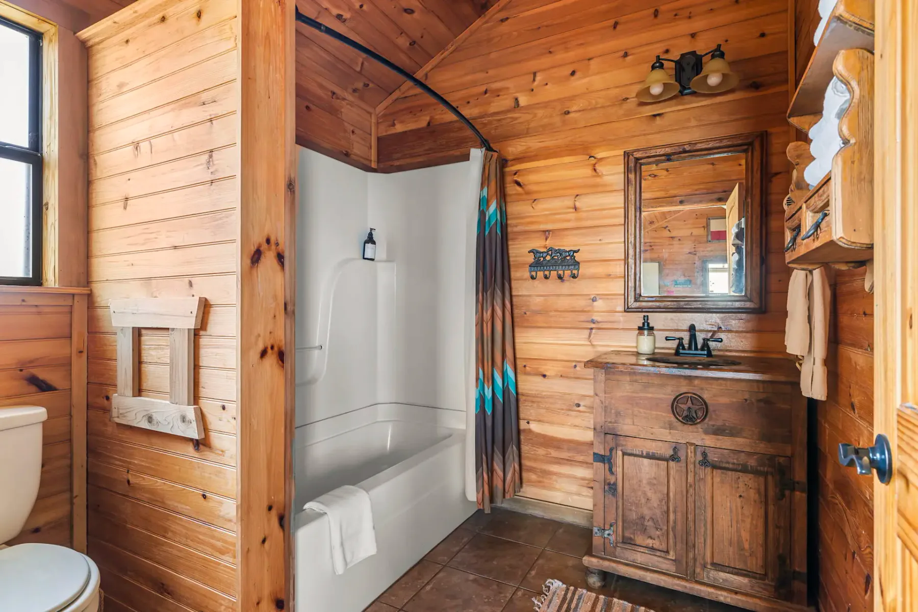 Baño rústico con revestimiento de madera, una bañera blanca, cortina de ducha de colores, mueble de baño de madera con espejo e inodoro.