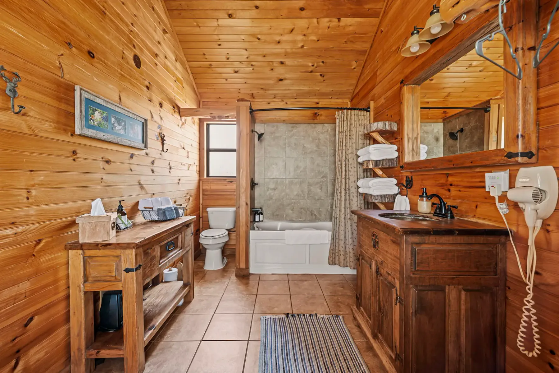 Baño revestido de madera con decoración rústica, inodoro, bañera con cortina, mueble de baño de madera, espejo, secador de pelo y objetos decorativos en una repisa.