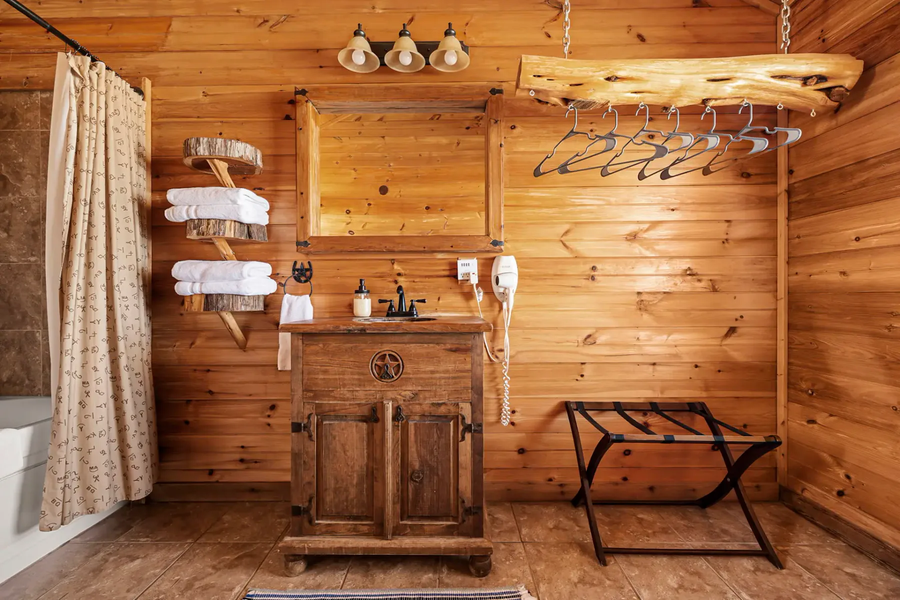 Baño rústico de madera con un mueble de baño de madera, toallas, ducha con cortina y una barra suspendida con ganchos para ropa junto a paredes revestidas de madera.