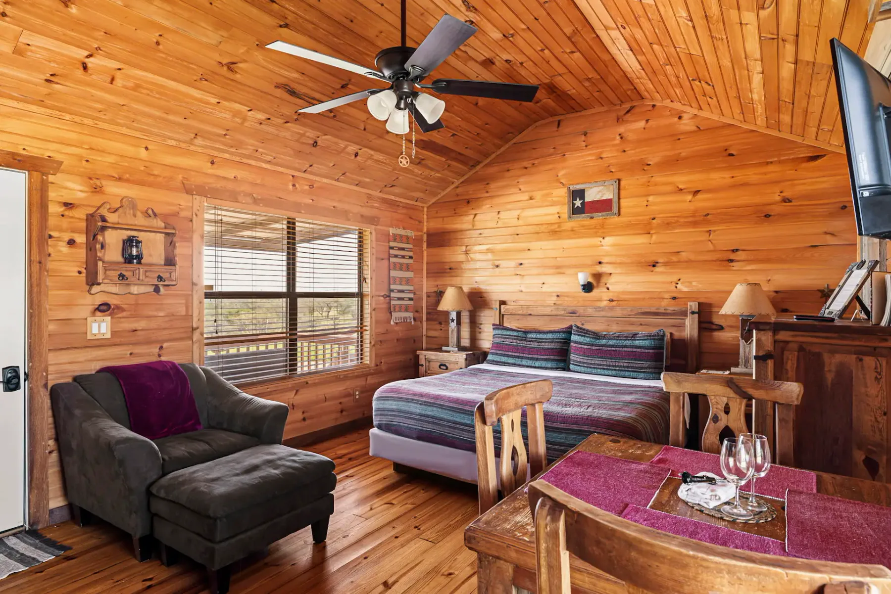 Acogedora habitación de cabaña con paredes de madera, una cama, un sillón, una pequeña mesa de comedor y un ventilador de techo, iluminada por la luz natural de una ventana.
