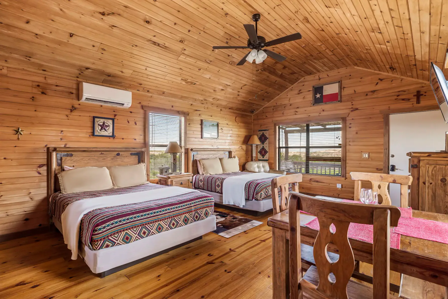 Una acogedora habitación de estilo cabaña con paredes y techo de madera, dos camas, un ventilador de techo y un pequeño comedor, adornada con decoración rústica.