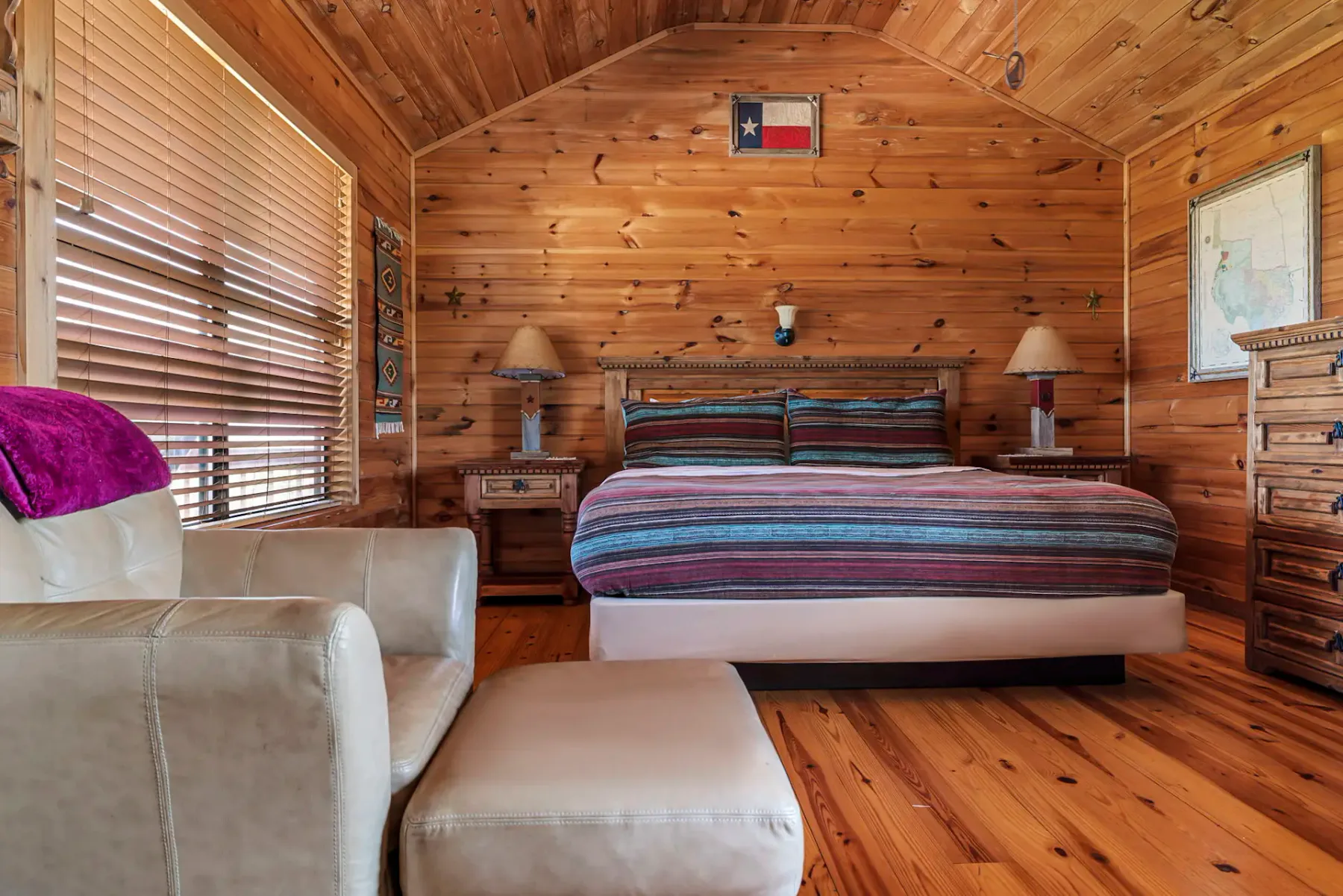 Dormitorio rústico de madera con una colcha de rayas, un sillón y un reposapiés. Una bandera de Texas en la pared, flanqueada por lámparas y una ventana con persianas.