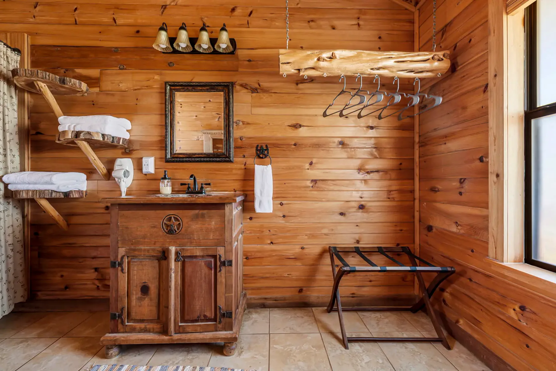 Baño rústico de madera con un pequeño mueble de baño, espejo, toalleros y perchero de estilo tronco. Entra luz natural por una ventana.
